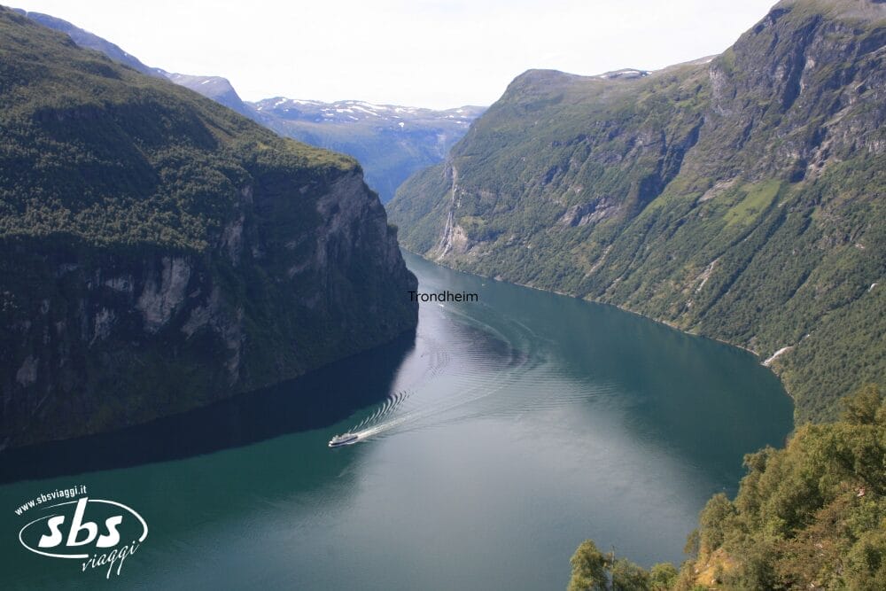 Una vista panoramica di un fiordo stretto e tortuoso in Norvegia, con montagne ripide e verdi su entrambi i lati. Un battello scivola nell'acqua blu e calma lungo la Via Atlantica. La scritta "Trondheim" appare vicino al centro; il logo "SBS Viaggi" è nell'angolo.