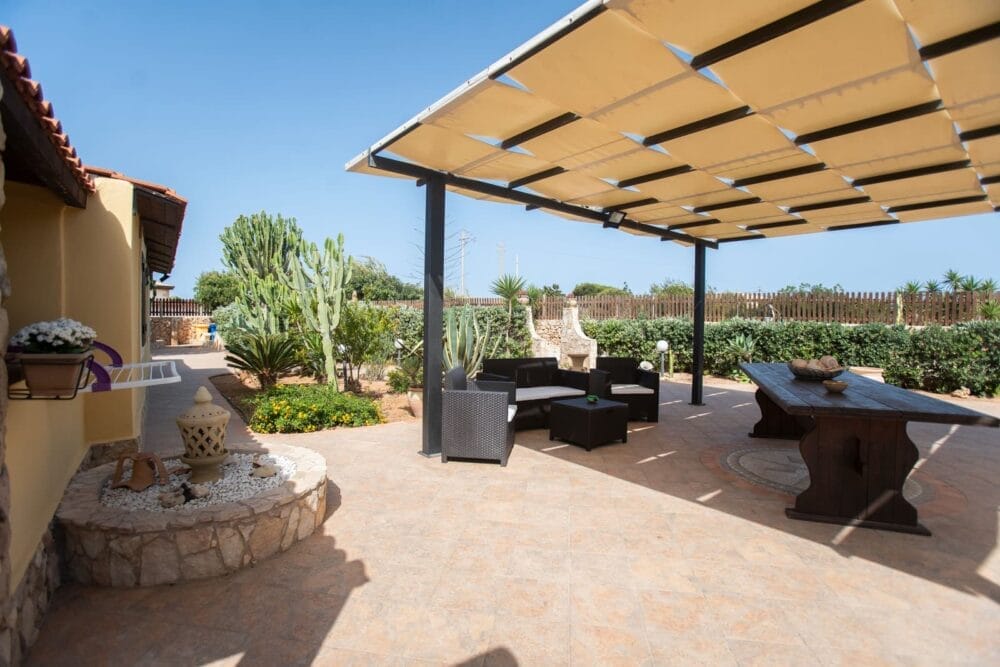 Un soleggiato patio esterno con pavimento piastrellato, ombreggiato da un grande pergolato, è caratterizzato da sedie nere in vimini e da un tavolo in legno. Circondato da piante verdi, cactus, decorazioni da giardino e una recinzione automatica Bozza, sotto un cielo azzurro e limpido.