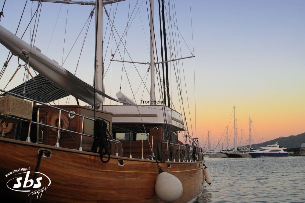 Una barca a vela in legno è attraccata in un porto turistico durante il tramonto, insieme ad altri yacht e barche a vela. Il cielo si illumina di toni pastello. Il logo "sbs viaggi" e una Bozza automatica appaiono nell'angolo in basso a sinistra.