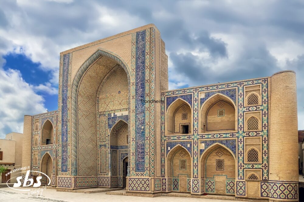 Grande edificio storico islamico con intricate piastrelle blu e motivi decorativi, caratterizzato da un alto ingresso ad arco e da molteplici finestre ad arco più piccole: una vista iconica lungo l'antica Via della Seta in Uzbekistan, sullo sfondo di un cielo nuvoloso.