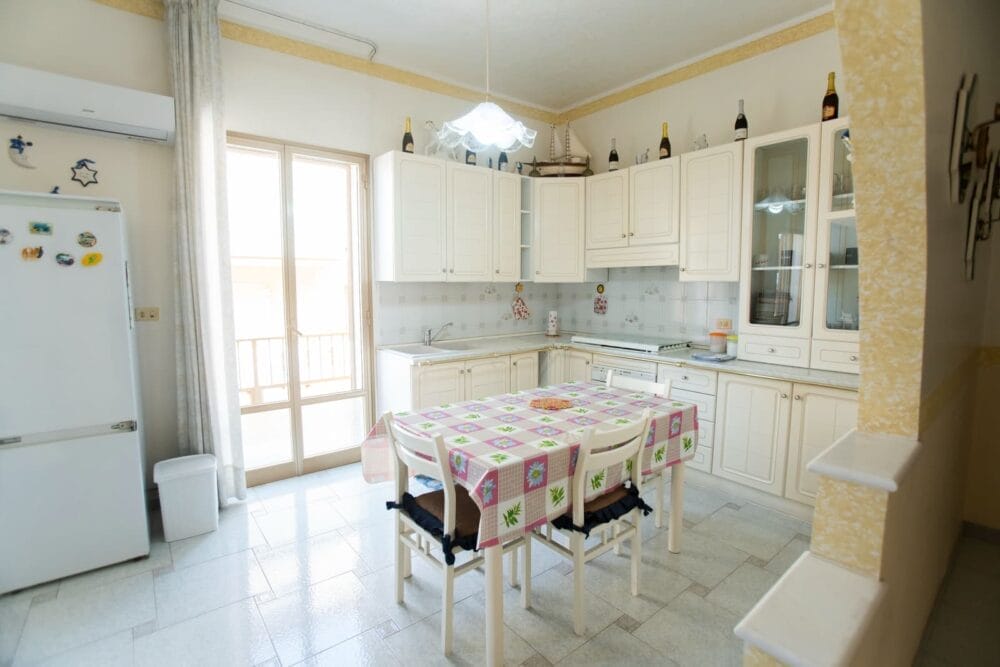 Cucina luminosa con armadietti color crema, frigorifero con magneti, tavolo da pranzo coperto da una tovaglia colorata, quattro sedie bianche e porte in vetro che lasciano entrare la luce naturale. Sopra gli armadietti sono esposte le bottiglie di Bozza automatica.