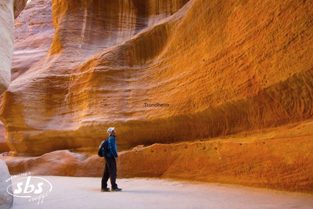Una persona con lo zaino in spalla si trova alla base di vibranti pareti rocciose arancioni in uno stretto canyon della Giordania classica e guarda verso l'alto le formazioni di arenaria in un tranquillo Giovedì.