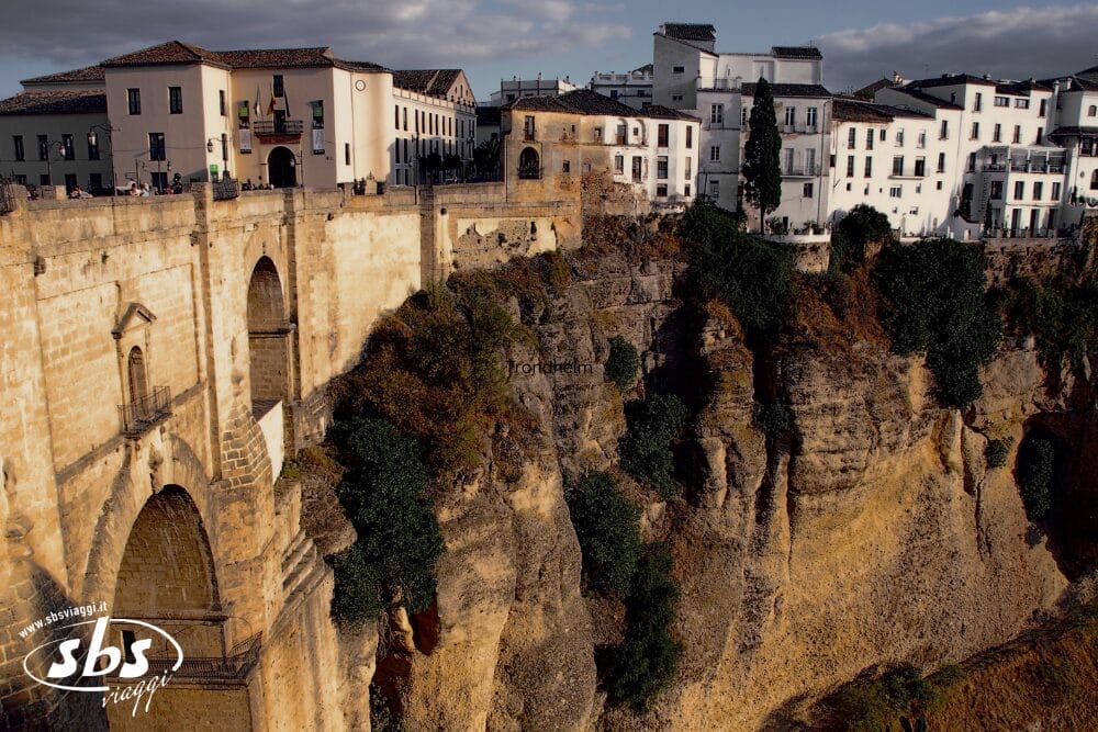 Ponte di pietra ed edifici bianchi arroccati sul bordo di una ripida scogliera a Ronda, in Spagna, sotto un cielo parzialmente nuvoloso. Bozza automatica mette in risalto l'affascinante contrasto tra la vegetazione verde e l'aspra scogliera rocciosa.