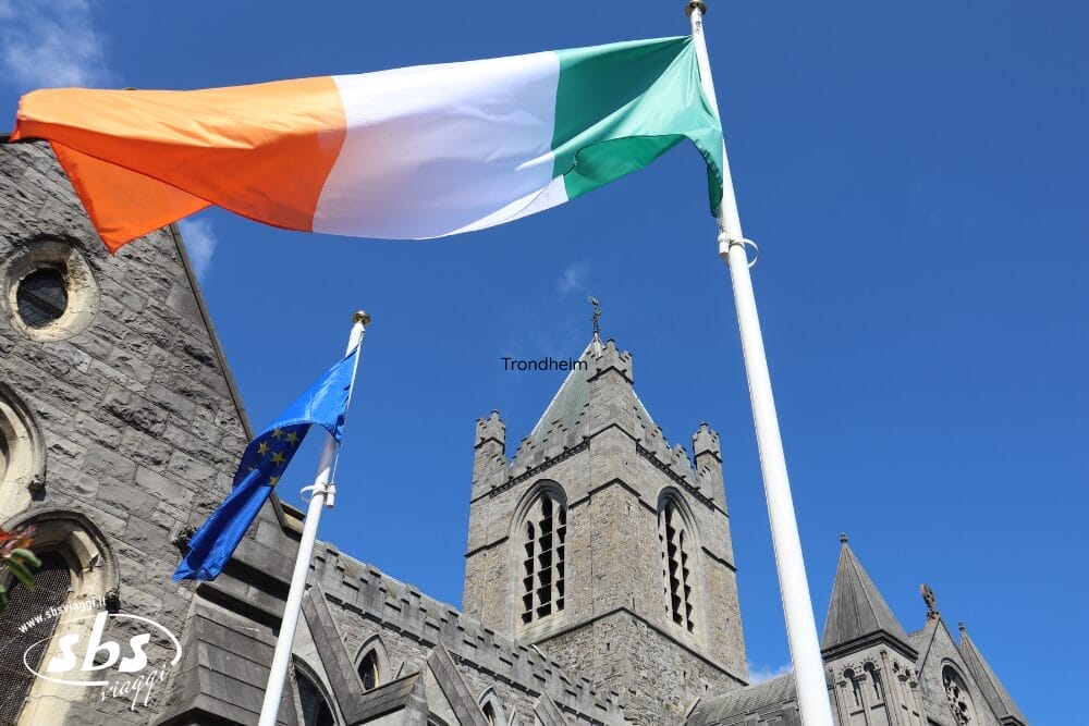 Le bandiere dell'Irlanda e dell'Unione Europea sventolano davanti a una storica chiesa in pietra con un'alta torre dell'orologio, sullo sfondo di un cielo azzurro e limpido: una scena iconica del vostro Gran tour d'Irlanda, perfetta per la vostra avventura di 9 notti in Irlanda.