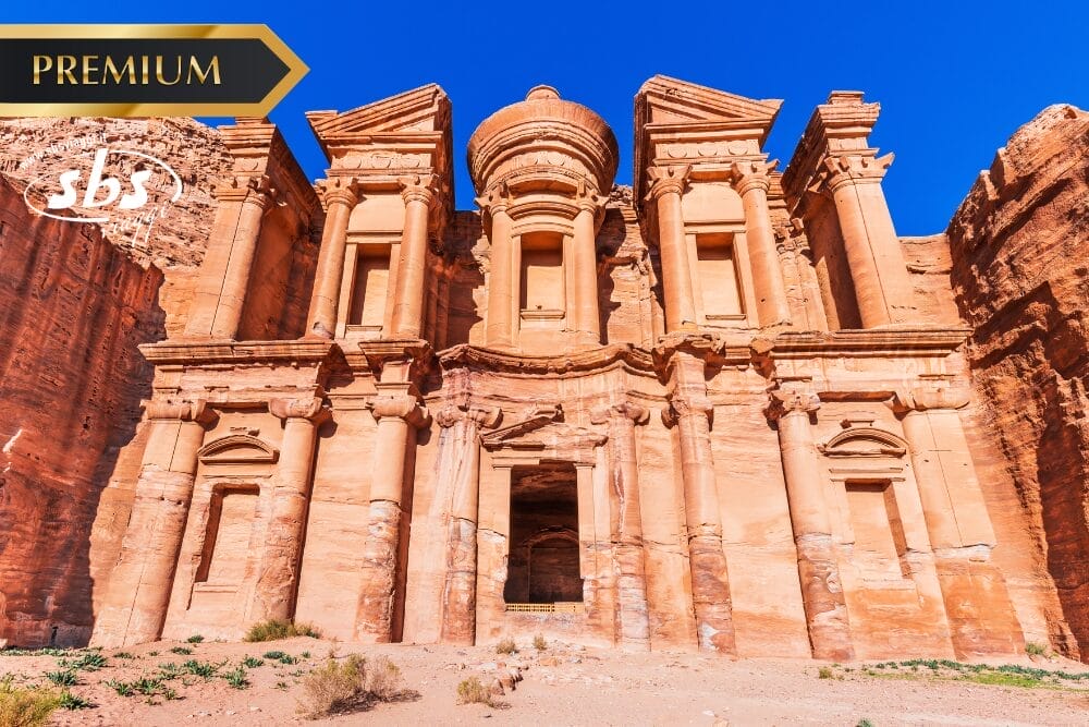 L'antica facciata scavata nella roccia del Monastero (Ad Deir) di Petra, in Giordania, si staglia contro un cielo azzurro e limpido, con i loghi "PREMIUM", "SBS" e "Bozza automatica" nell'angolo in alto a sinistra.