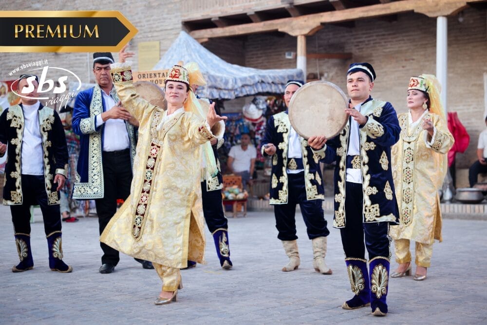 Un gruppo di persone in abiti tradizionali dell'Asia centrale si esibisce in una vivace danza popolare all'aperto. Una persona suona un tamburo a cornice, mentre altre in abiti e cappelli ornati danzano gioiosamente. Sullo sfondo, gli spettatori assistono alla vibrante celebrazione della Bozza automatica.