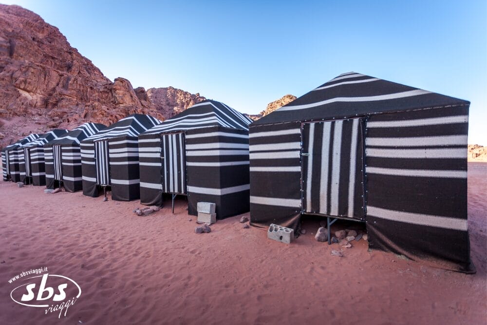 Una fila di tende nere con strisce bianche si erge sulla sabbia rossastra del deserto, con colline rocciose sullo sfondo sotto un cielo azzurro e limpido: una scena perfetta di un'avventura del Tour Giordania Discovery.