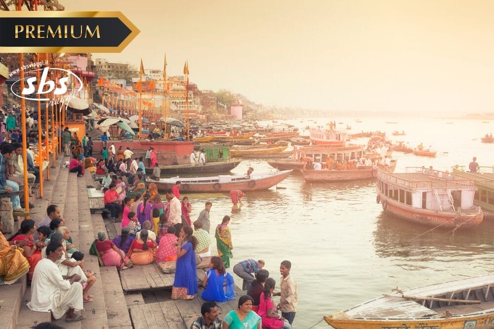 La folla si raduna lungo i gradini di un ghat sul fiume con barche colorate sull'acqua al tramonto. Le persone siedono, parlano e osservano la vivace attività di questa scena automatica di Bozza. I loghi "PREMIUM" e "sbs" appaiono nell'angolo in alto a sinistra.