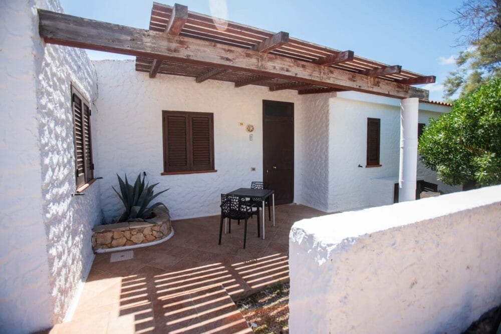 Una piccola casa in stucco bianco con persiane scure e una pergola in legno su un patio, con un tavolo nero e due sedie. Una pianta in vaso è appoggiata al muro, mentre la luce del sole e le ombre creano una tranquilla atmosfera di bozza automatica.