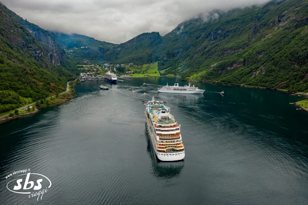 Veduta aerea di alcune navi da crociera che attraversano i fiordi della Norvegia lungo la panoramica Via Atlantica, circondate da verdi montagne e da un piccolo villaggio, con l'acqua calma che riflette lo splendido scenario.