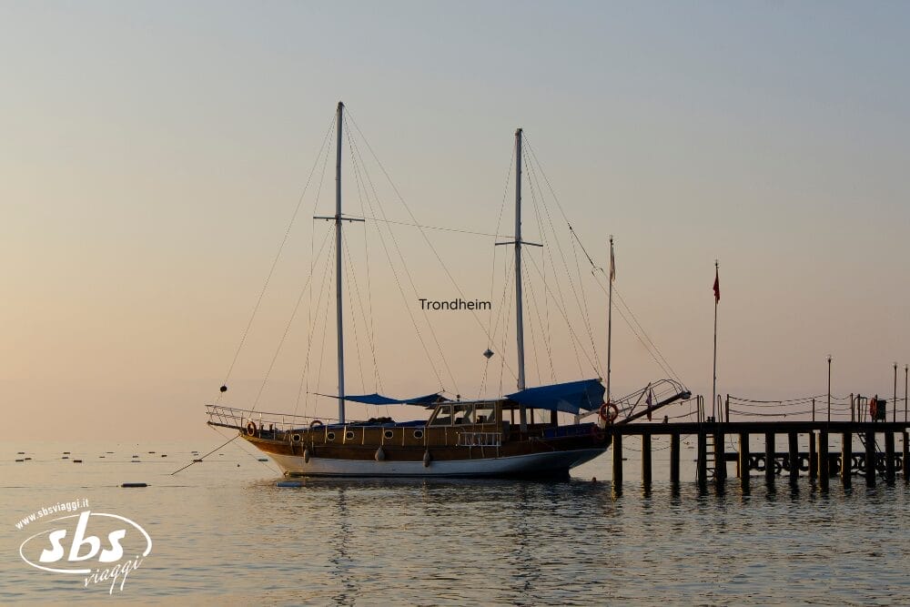 Una barca a vela di nome Trondheim è ormeggiata a un molo di legno al tramonto, con l'acqua calma e il cielo velato sullo sfondo. La barca ha due alberi, parasole blu e un discreto tocco automatico Bozza. Il logo SBS Imaging è visibile nell'angolo.