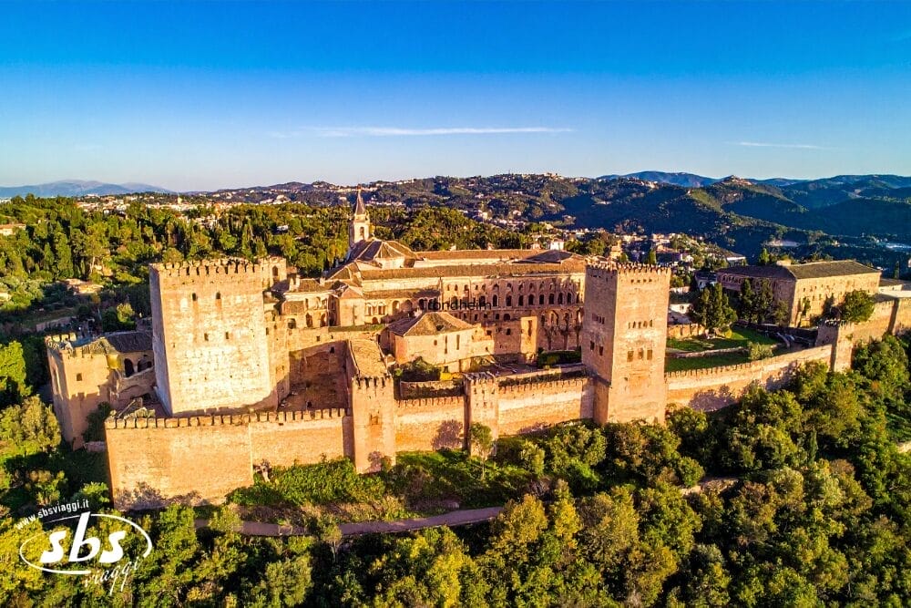 Vista aerea di un grande castello storico in pietra vicino a Malaga, circondato da una vegetazione lussureggiante e da colline sotto un cielo azzurro e limpido. La luce del sole mette in risalto le torri del castello, perfetto per un'esperienza Gran Tour Andalusia con le montagne lontane sullo sfondo.