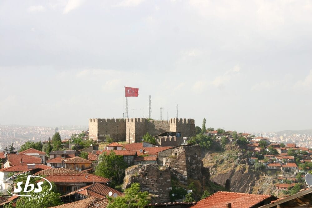 Una fortezza in pietra con una grande bandiera turca si erge su una collina circondata da case dai tetti rossi ad Ankara, in Turchia, sotto un cielo parzialmente nuvoloso: una scena classica che ricorda un tour turco di Bodrum.