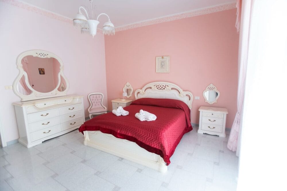 Una camera da letto con mobili bianchi, tra cui un letto con copriletto rosso, due comodini, un comò con specchio e cigni di spugna sul letto. Le pareti rosa e il pavimento grigio chiaro creano un ambiente accogliente e automatico.