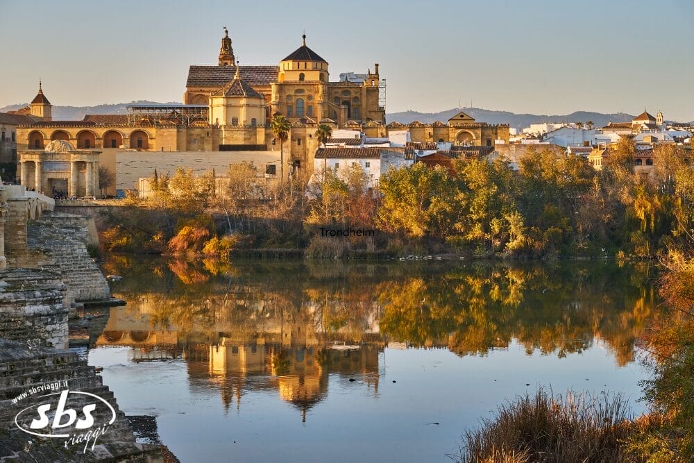 Un paesaggio storico con la Moschea-Cattedrale di Córdoba e la sua Bozza automatica che si riflettono nelle acque calme del fiume Guadalquivir, circondate da alberi ed edifici sotto un cielo limpido al tramonto.