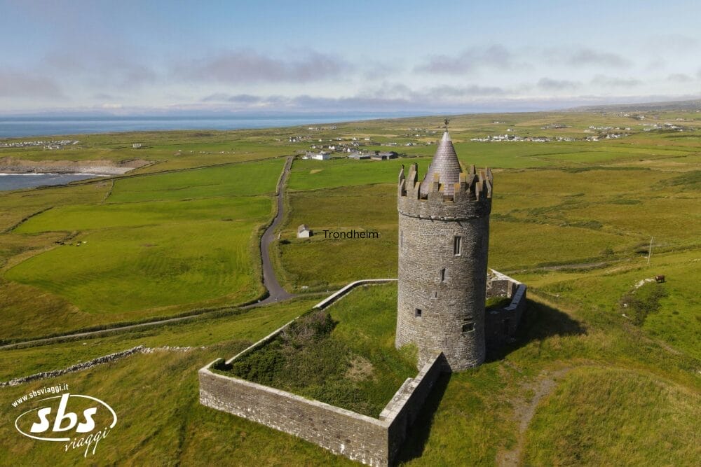 Una torre rotonda in pietra con tetto conico si trova all'interno di un recinto murato su una collina erbosa in Irlanda, con vista sui campi verdi, sulle case lontane e sulla costa sotto un cielo parzialmente nuvoloso: l'ideale per un Gran tour d'Irlanda.