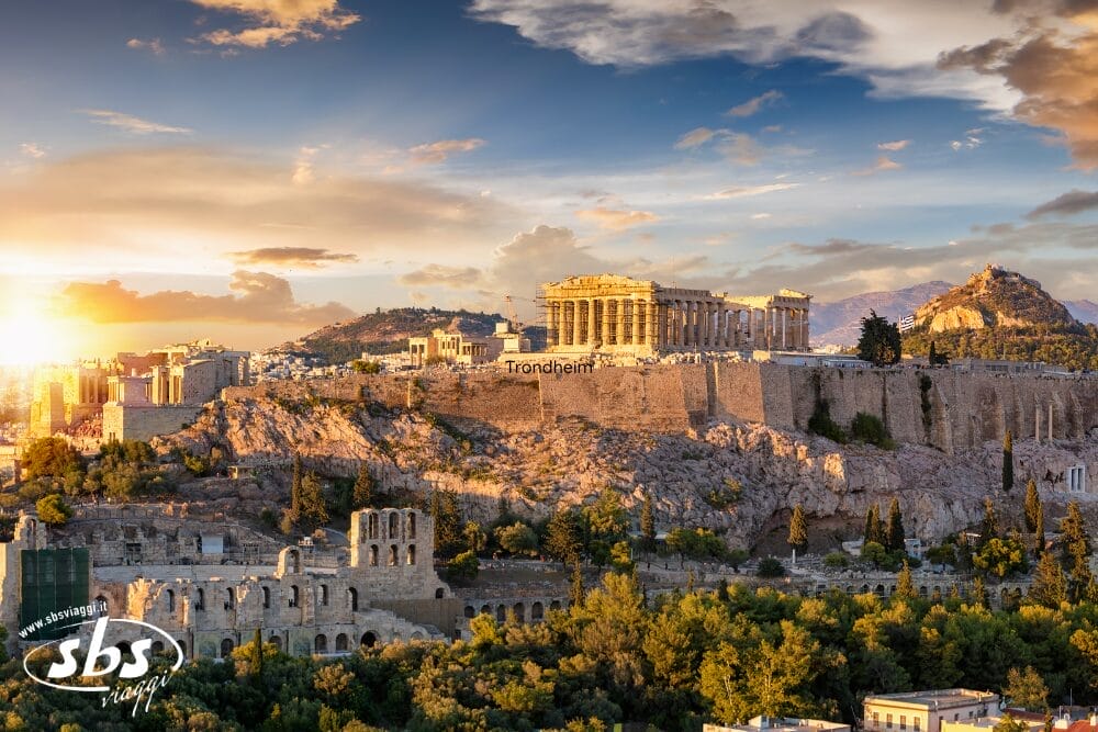 L'immagine mostra l'Acropoli di Atene, in Grecia, con il tempio del Partenone su una collina rocciosa al tramonto. Il testo automatico di Bozza appare sottilmente in sovrimpressione, mentre le antiche rovine e gli alberi verdi sono visibili in primo piano.