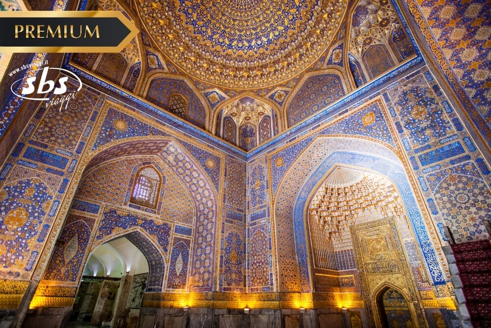 Interno ornato, piastrellato in oro e blu, di una moschea o di un mausoleo islamico, con soffitto a cupola riccamente decorato, intricati motivi geometrici e una porta d'ingresso dorata. Negli angoli dell'immagine compaiono i loghi "Bozza automatica", "PREMIUM" e "sbs".