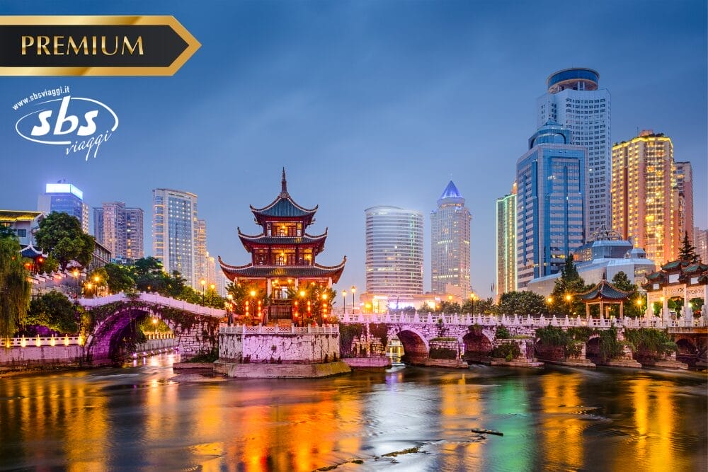 Una pagoda tradizionale e un ponte di pietra ad arco risplendono al tramonto, sullo sfondo del moderno skyline di Guizhou. Le parole "PREMIUM", "sbs viaggi" e "Bozza automatica" compaiono nell'immagine.