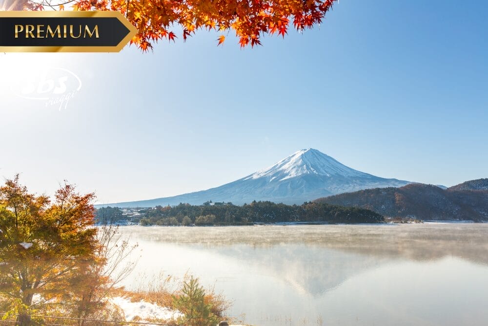 Veduta del Monte Fuji con la cima innevata, il cielo azzurro e le foglie autunnali in primo piano, riflessa in un lago calmo con una leggera foschia. L'etichetta "Premium" e Bozza automatica appaiono nell'angolo in alto a sinistra.