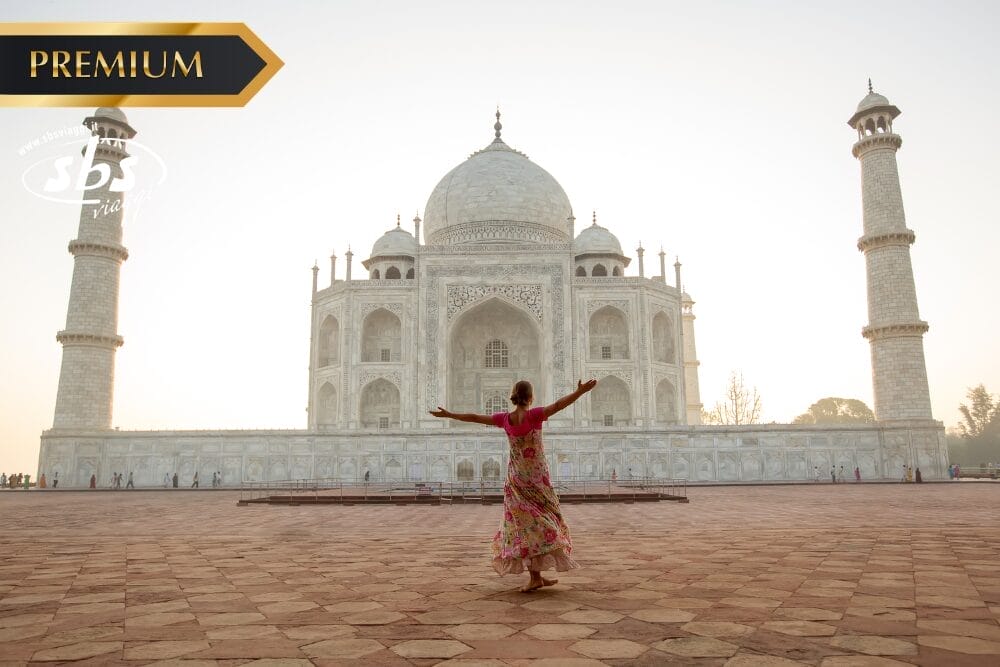 Una persona in abiti colorati sta in piedi con le braccia aperte davanti al Taj Mahal all'alba, in un ampio cortile di pietra rossa. Nell'angolo in alto a sinistra si trova la scritta "PREMIUM", a sinistra un logo bianco e sotto, in modo discreto, la scritta "Bozza automatica".