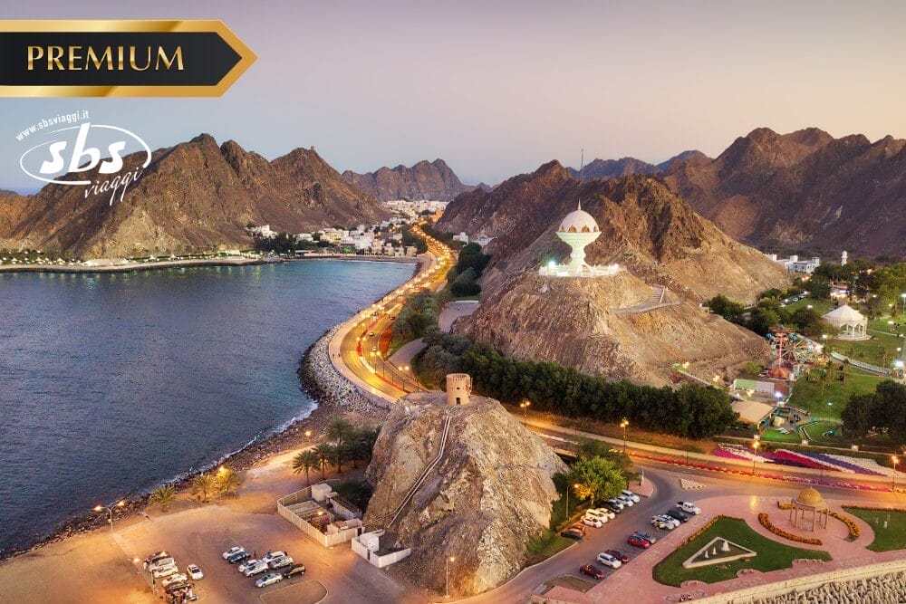 Paesaggio urbano costiero al tramonto con strade tortuose, montagne rocciose, un monumento bianco su una collina, un parco e auto parcheggiate. La scritta "Bozza automatica" e i loghi "PREMIUM" e "sbs viaggio" appaiono in alto, mentre il mare delimita la città.