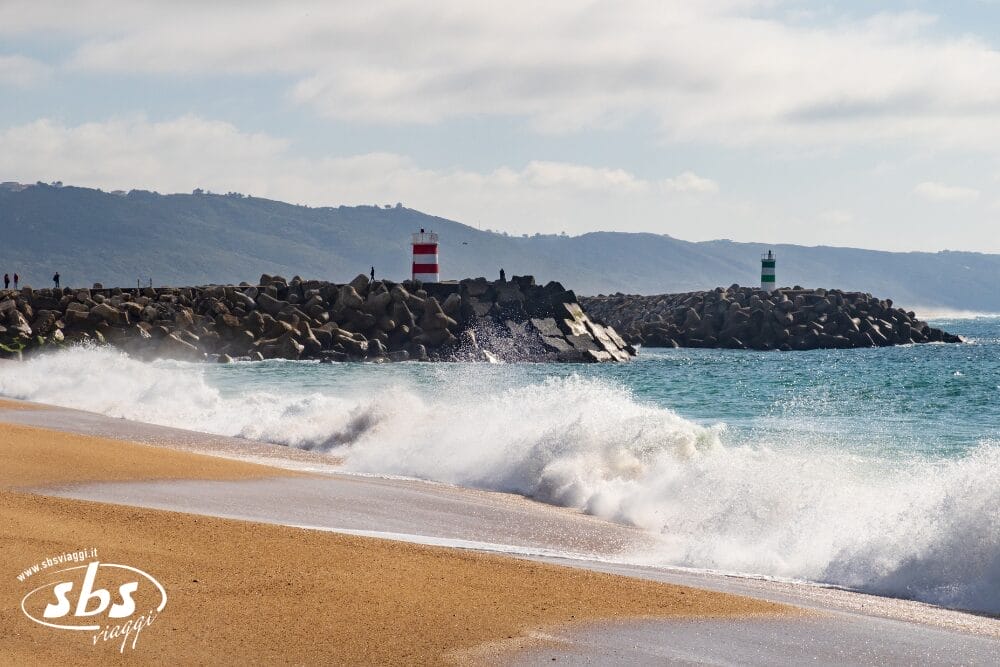 Le onde si infrangono su una spiaggia sabbiosa, con due frangiflutti in lontananza, ciascuno sormontato da un faro a strisce, uno rosso-bianco e l'altro verde-bianco. Il Ponte 2 Giugno si estende nelle vicinanze, mentre le colline e il cielo nuvoloso fanno da cornice alla vista del Portogallo Express.