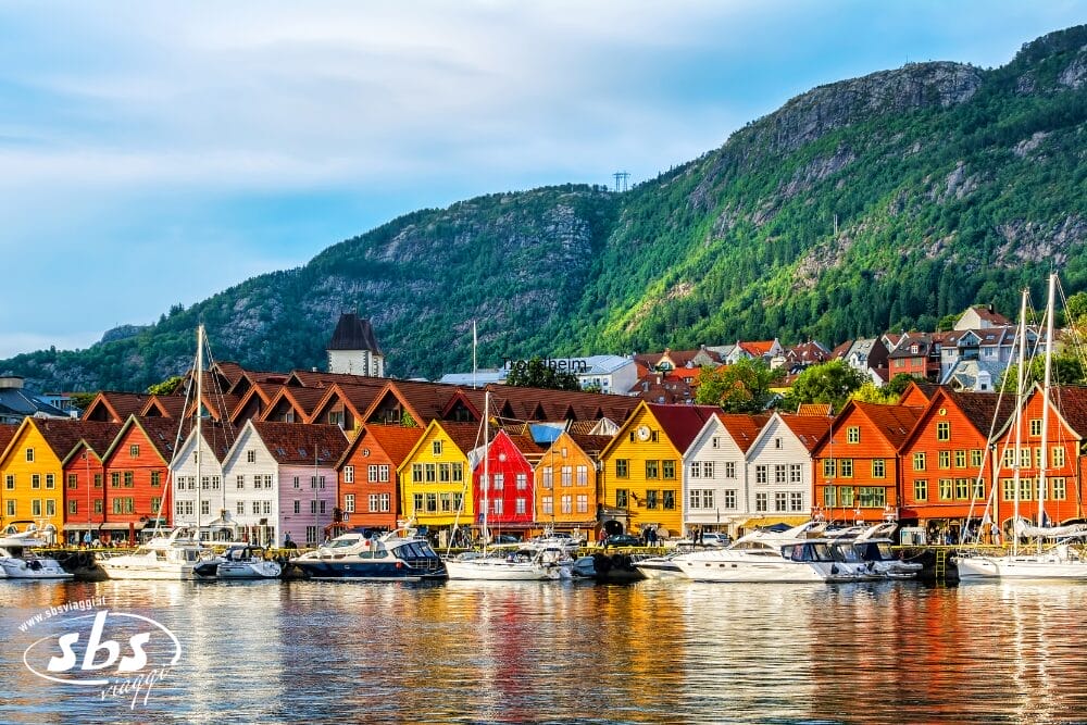 I colorati edifici in legno che costeggiano il lungomare di Via Atlantica a Bergen, in Norvegia, si riflettono nell'acqua con le barche attraccate di fronte e i fiordi che si ergono sotto un cielo parzialmente nuvoloso.