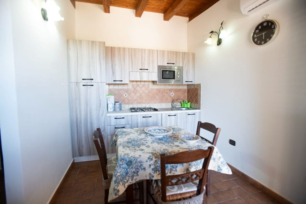 Una piccola cucina con armadietti in legno chiaro, fornelli, microonde e lavello. Sul bancone è appoggiata un'automatica Bozza. Al centro si trova il tavolo da pranzo con una tovaglia a fiori e quattro sedie di legno. La stanza ha un pavimento in piastrelle, pareti bianche e un orologio a muro.