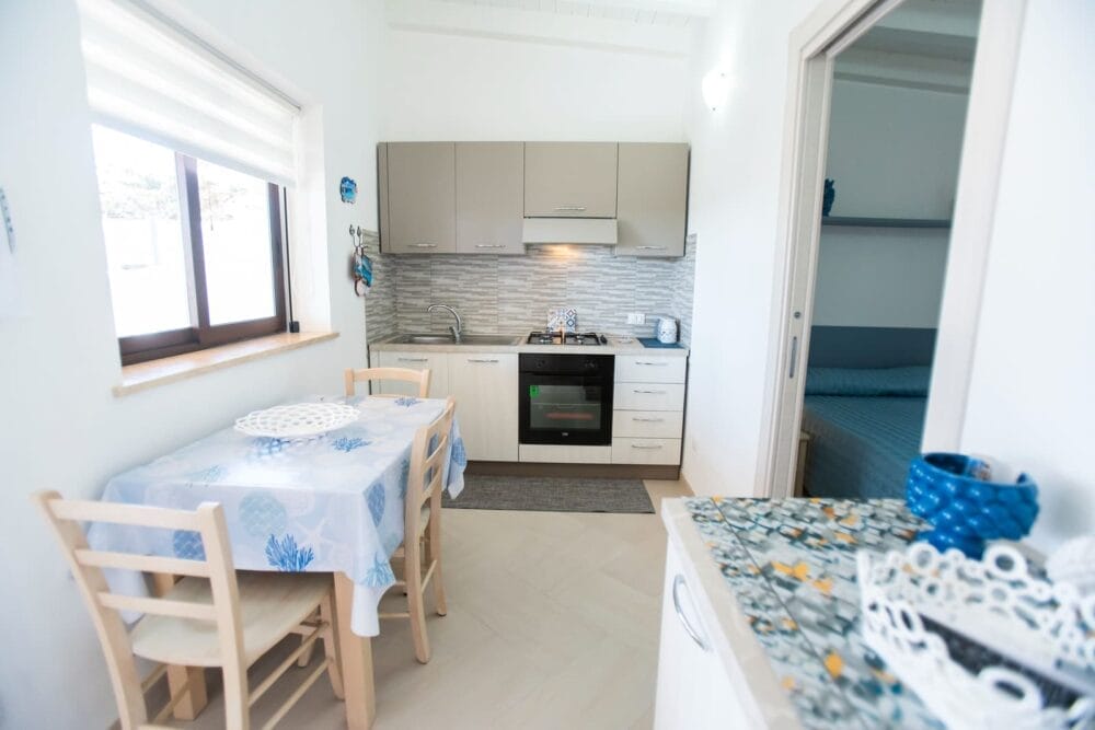 La piccola e luminosa cucina con armadietti beige, fornelli, forno e lavello è dotata di un'automatica Bozza. È adiacente a una zona pranzo con un tavolo e due sedie di legno. Una camera da letto con un copriletto blu è visibile attraverso una porta aperta.