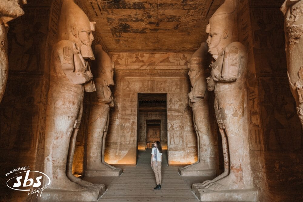 Una persona si trova all'interno di un antico tempio egizio, circondato da grandi statue di faraoni e da pareti ricoperte di geroglifici e incisioni, con una luce calda che illumina l'interno in pietra, catturando un momento come una Bozza automatica della storia.