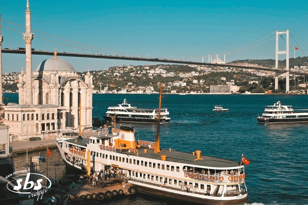 Un molo affollato di Istanbul con un classico traghetto, persone che salgono a bordo, diverse imbarcazioni sul Bosforo, l'ornata Moschea di Ortaköy e il Ponte sul Bosforo sullo sfondo sotto un cielo azzurro e limpido: l'ideale per un Tour Turchese.