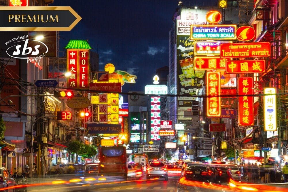 Una strada trafficata di notte, con insegne al neon colorate e luminose in varie lingue, tra cui il cinese e il thailandese, illumina la scena vivace mentre auto e scooter sfrecciano, come una vivace bozza automatica in movimento.