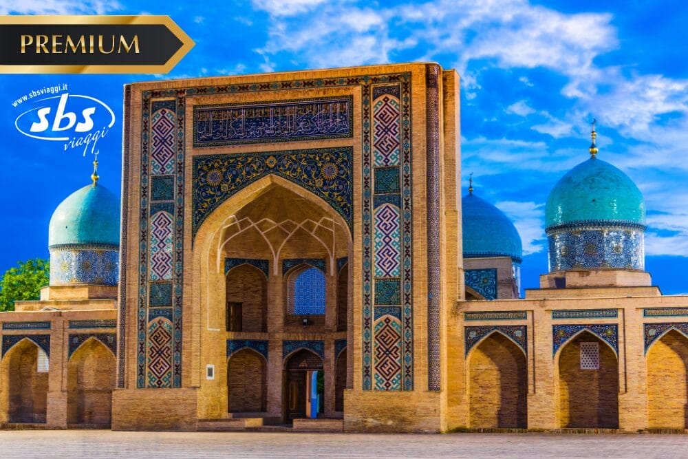 Una grande moschea con intricate piastrelle, un alto ingresso ad arco e cupole blu sotto un cielo azzurro brillante. I loghi "PREMIUM" e "sbs viaggi", insieme all'etichetta Bozza automatica, sono visibili negli angoli superiori dell'immagine.