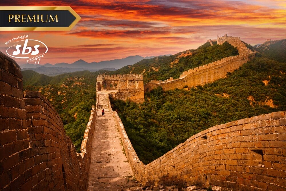 Una vista panoramica della Grande Muraglia Cinese si estende su verdi colline al tramonto, con nuvole arancioni e vivaci in alto. Due persone camminano lungo la muraglia e nell'angolo compare un'etichetta di Bozza automatica "PREMIUM" con il logo di SBS Viaggi.