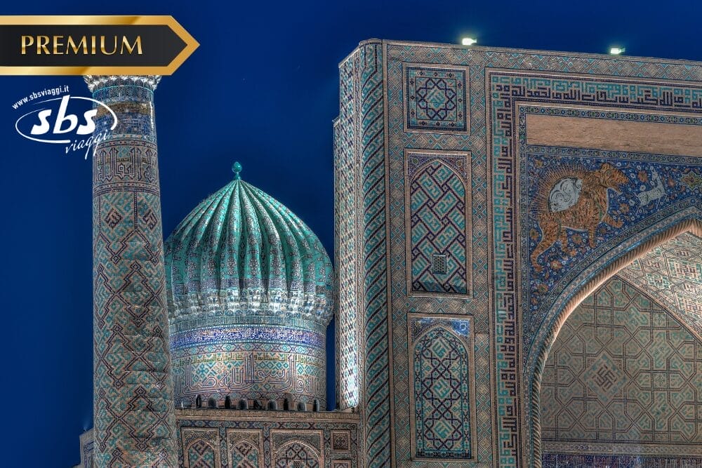 Veduta notturna di un edificio islamico con tetto a cupola e minareto, decorato in modo intricato e con piastrelle blu, con motivi geometrici ornamentali. L'immagine include i loghi "PREMIUM", "sbs viaggi" e una sottile sovrapposizione di Bozza automatica nell'angolo.