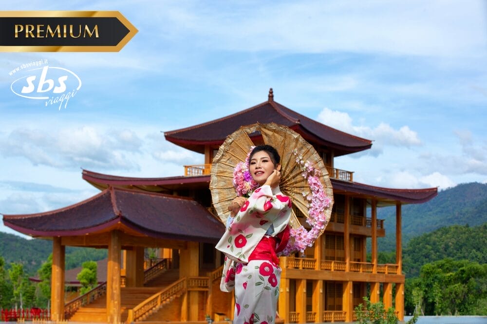 Una donna con un kimono floreale regge un ombrellino decorativo e sorride davanti a un grande edificio in legno con i tetti curvi - che cattura il fascino di Bozza automatica - incastonato tra le colline verdi e il cielo azzurro.