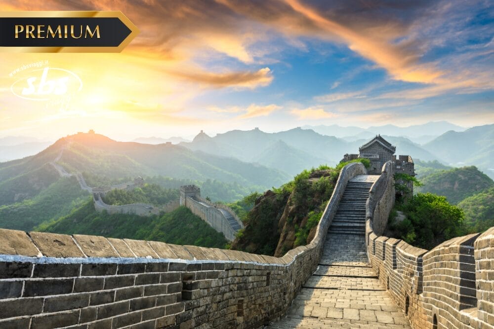 Un sentiero di pietra della Grande Muraglia Cinese si estende in lontananza su verdi colline al tramonto, con la scritta "PREMIUM", un debole logo "sbs" e una sottile filigrana bozza automatica nell'angolo superiore sinistro.