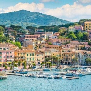 Colorato villaggio di mare con edifici color pastello, barche attraccate in un porticciolo e colline verdi sullo sfondo sotto un cielo parzialmente nuvoloso.