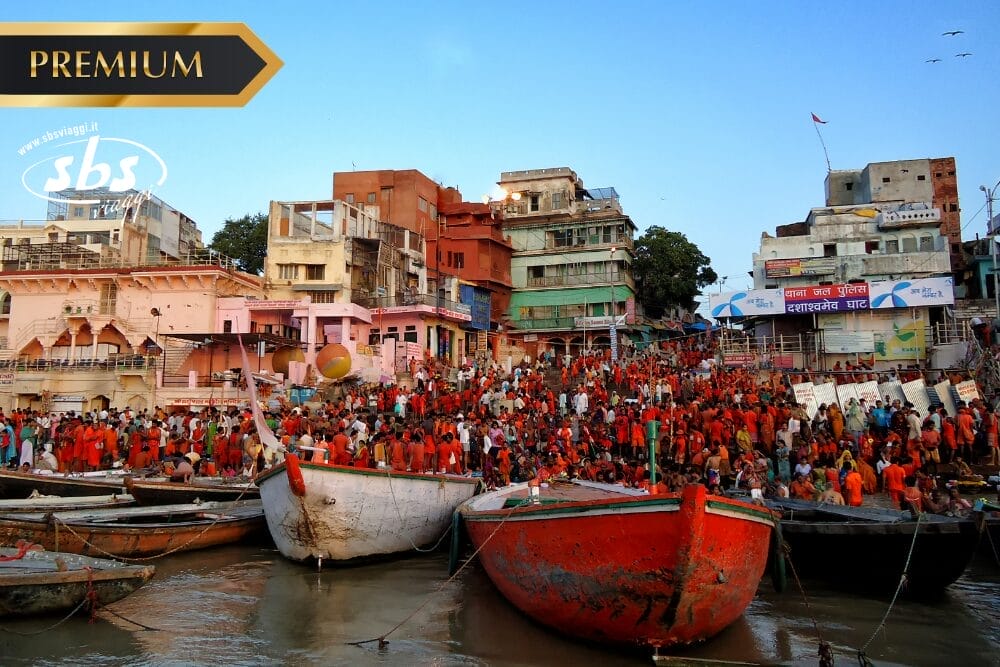 Folla di persone che si raduna sui ghat lungo il fiume a Varanasi, in India, con edifici colorati e barche lungo l'acqua durante il giorno; etichette "Bozza automatica" e "PREMIUM" in alto a sinistra e filigrana "sbs" sull'immagine.