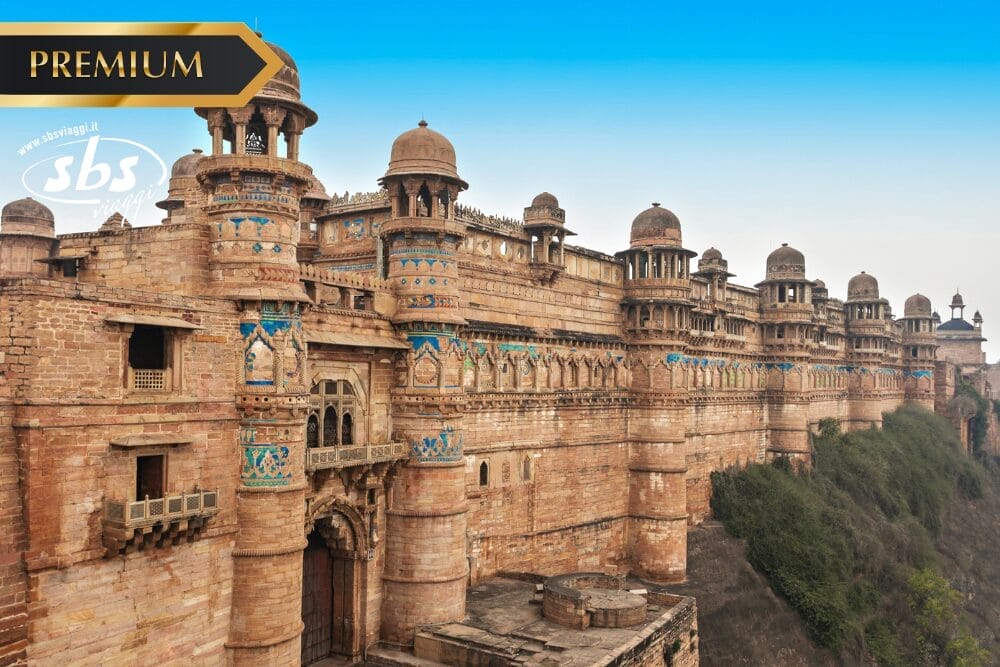 Una vista panoramica dello storico Forte di Gwalior in India, caratterizzato da pareti in pietra decorate, torri a cupola e accenti di piastrelle blu sotto un cielo limpido. I loghi "PREMIUM", "SBS" e "Bozza automatica" sono visibili nell'angolo superiore sinistro.