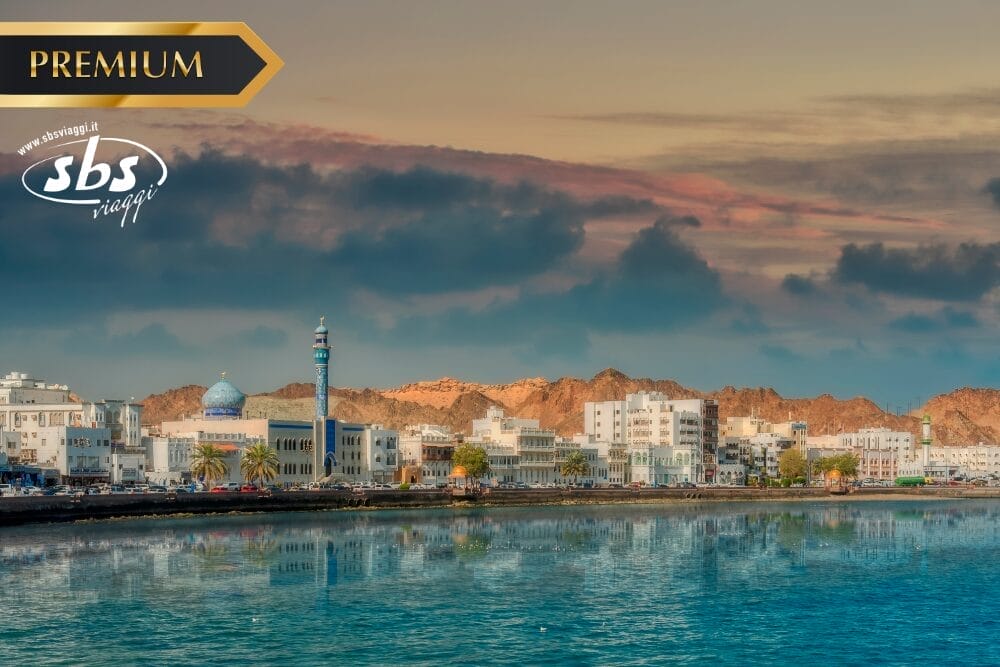 Una città panoramica sul lungomare con edifici bianchi, una moschea dalle cupole blu, palme e montagne sullo sfondo sotto un cielo colorato, con i loghi "PREMIUM", "sbs viaggi" e Bozza automatica travel nell'angolo.