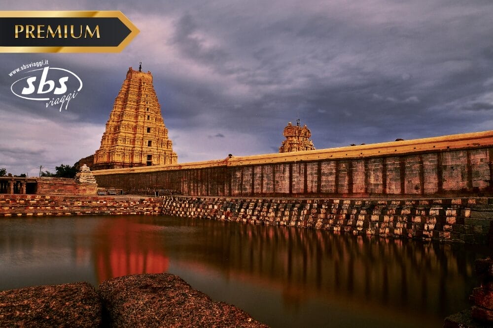 Un grande tempio in pietra con intricati intagli si trova accanto a un serbatoio d'acqua riflettente sotto un drammatico cielo nuvoloso. I loghi "PREMIUM", "sbs viaggi" e "Bozza automatica" appaiono nell'angolo superiore sinistro dell'immagine.