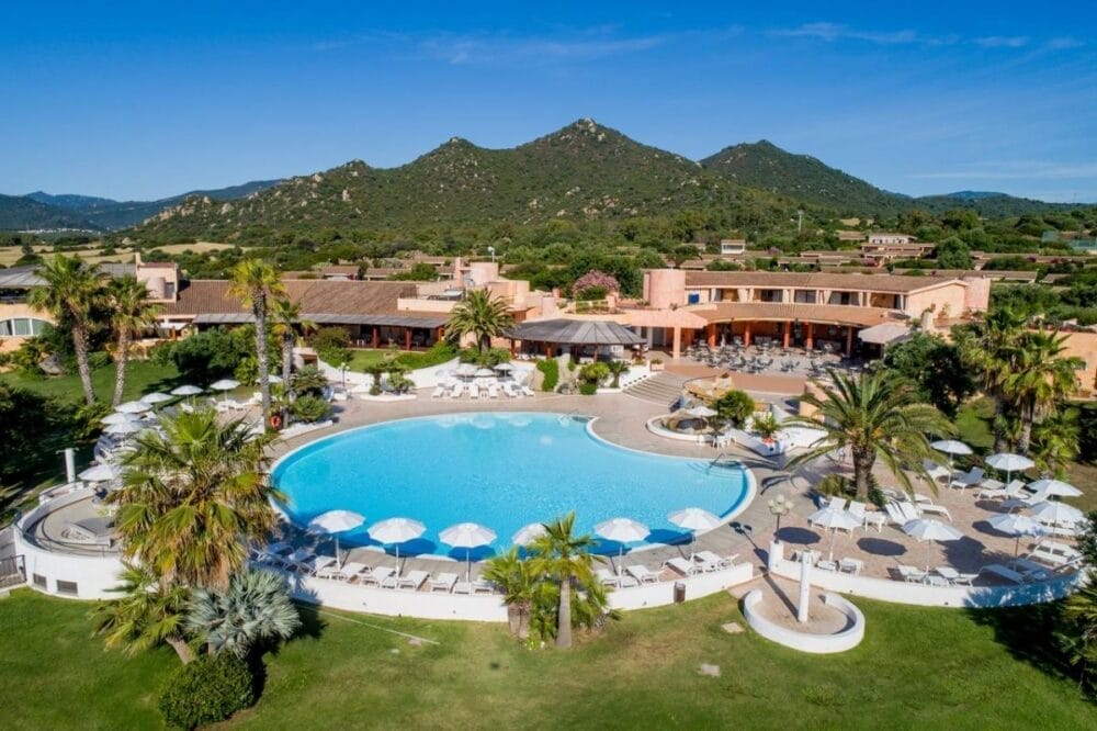 Veduta aerea di un resort con una grande piscina curva circondata da palme, sedie a sdraio e ombrelloni bianchi, sullo sfondo di colline verdi e di un cielo azzurro e limpido: l'ideale per rilassarsi o per preparare la prossima Bozza automatica.