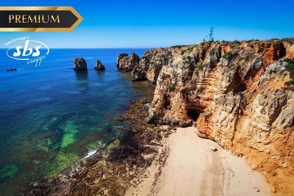 Una vista panoramica di scogliere e formazioni rocciose lungo una costa azzurra e trasparente, con una spiaggia sabbiosa sottostante. La scritta "Bozza automatica" appare vicino ai loghi SBS Viaggi e PREMIUM nell'angolo in alto a sinistra.