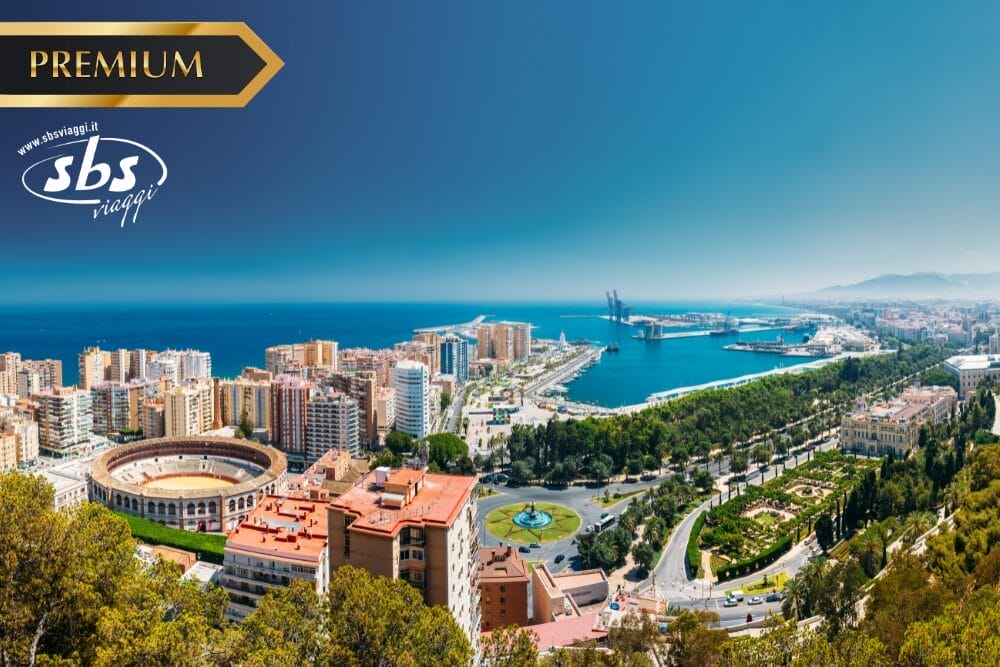 Vista panoramica di una città costiera con un'arena, alti edifici, parchi verdi e un porto blu sotto un cielo limpido. I loghi "PREMIUM", "SBS Viaggi" e "Bozza automatica" sono visualizzati nell'angolo in alto a sinistra.