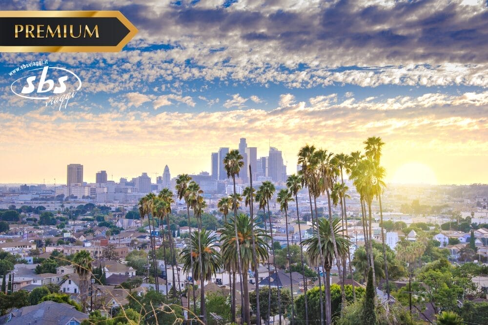 Una vista panoramica del centro di Los Angeles all'alba con alte palme in primo piano, edifici cittadini sullo sfondo e un cielo parzialmente nuvoloso. La scritta "PREMIUM", il logo SBS e "Bozza automatica" sono sovrapposti negli angoli superiori.
