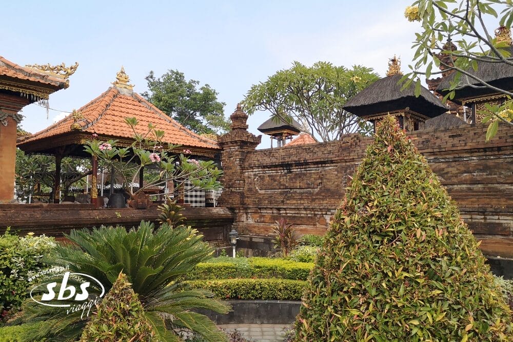 Architettura tradizionale balinese a Seminyak, con tetti di tegole rosse, muri di mattoni decorati, cespugli verdi e ordinati in un giardino sereno sotto il cielo azzurro di Bali.
