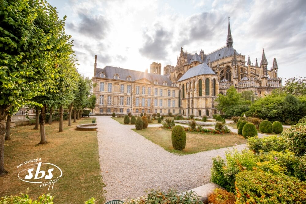 Veduta grandangolare di una storica cattedrale gotica in una delle Capitali d'Europa, con alte guglie e finestre ad arco, accanto a un giardino formale con cespugli curati, alberi e sentieri di ghiaia sotto un cielo parzialmente nuvoloso.