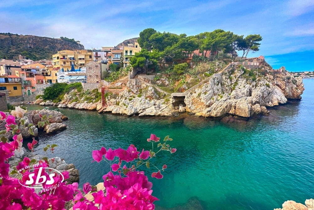 Un villaggio costiero con edifici colorati si trova in cima a scogliere rocciose accanto a un'acqua turchese e trasparente, circondato da una vegetazione lussureggiante e da vivaci fiori rosa in primo piano: un luogo perfetto per immortalare una Bozza automatica.